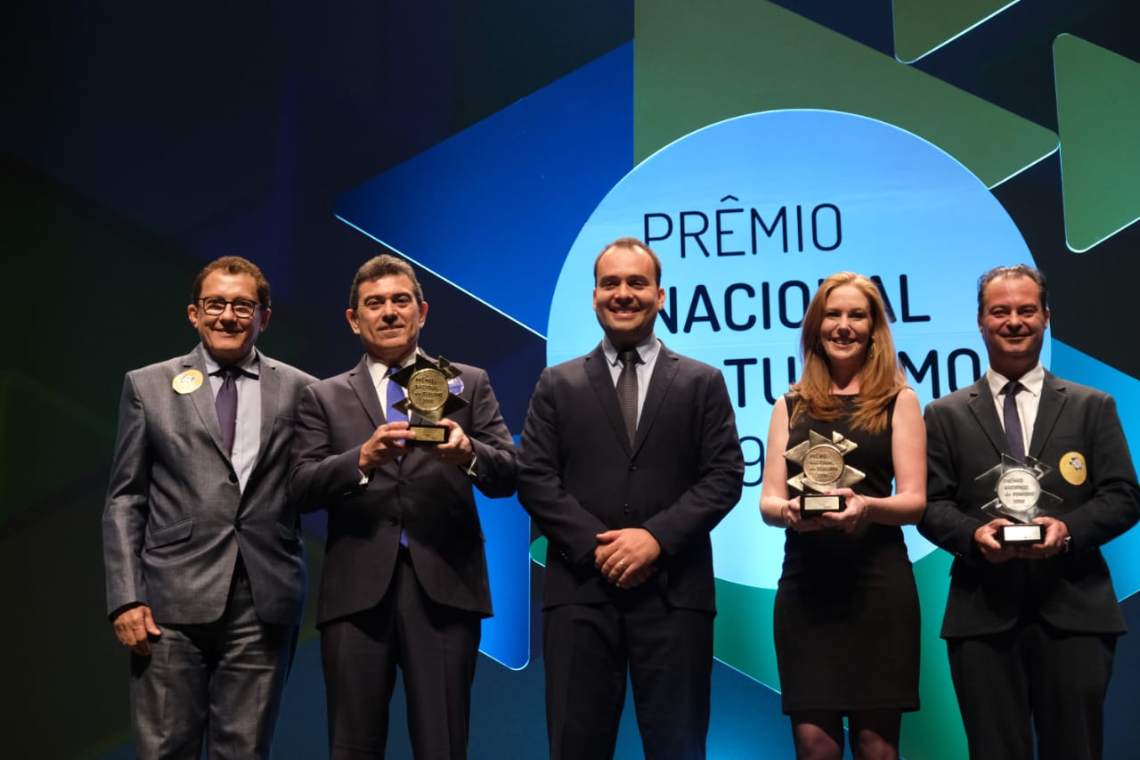 secretário em pé segurando prêmio ao lado de pessoas em cima de palco 